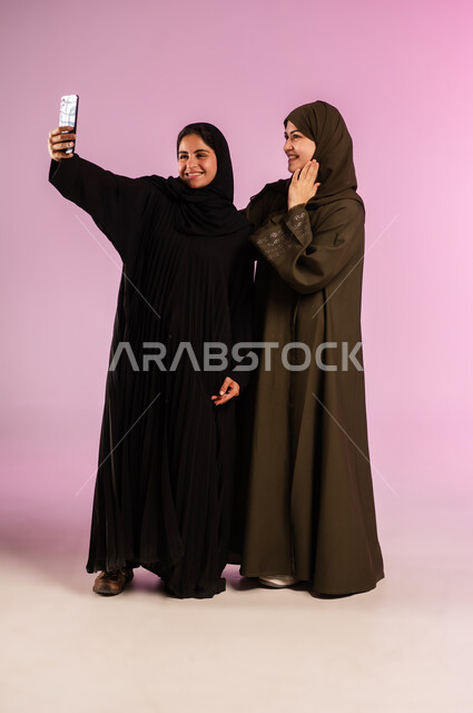 توثيق اللحظات السعيدة، استخدام الأجهزة التقنية المتطورة، بورتريه لشابة عربية خليجية سعودية محجبة ترتدي العباءة والحجاب تقوم بالتقاط صور السيلفي التذكارية مع صديقتها بإيماءات السعادة والسرور، صورة الجسم بالكامل، خلفية ملونة