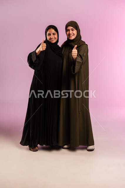 الاتفاق على أمر معين، شابة تعانق اختها، القبول والرضا بالنتيجة، ايماءات الاعجاب والموافقة،  بورتريه لإمرأتين عربيتين خليجيتين سعوديتين ترتديان الحجاب والعباءة ترفعان اصبع الابهام للأعلى بعلامة الجودة، صورة لكامل الجسم، خلفية ملونة