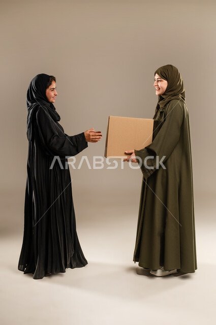 خدمات التوصيل المنزلي، استلام الطلبات الالكترونية، بورتريه لإمرأتين عربيتين خليجيتين سعوديتين ترتديان الحجاب والعباءة تحملان صندوق مشتريات الاونلاين السريع، جودة شركات الشحن السعودية، صورة الجسم بالطول الكامل، خلفية رمادية