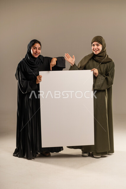 استخدام لوحات توضيحية ووسائل تعليمية، لوح اعلاني فارغ، الإشارة بإصبع السبابة بإيماءات السرور والاعجاب، إعلانات وعروض تسويقية، بورتريه لإمرأتين عربيتين خليجيتين سعوديتين ترتديان الحجاب والعباءة تمسكان موك اب مستطيل كبير، الجسم بالطول الكامل، خلفية رمادية