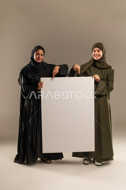 لوح اعلاني فارغ، الإشارة بإصبع السبابة بإيماءات السرور والاعجاب، استخدام لوحات توضيحية ووسائل تعليمية، إعلانات وعروض تسويقية، بورتريه لإمرأتين عربيتين خليجيتين سعوديتين ترتديان الحجاب والعباءة تمسكان موك اب مستطيل كبير، الجسم بالطول الكامل، خلفية رمادية