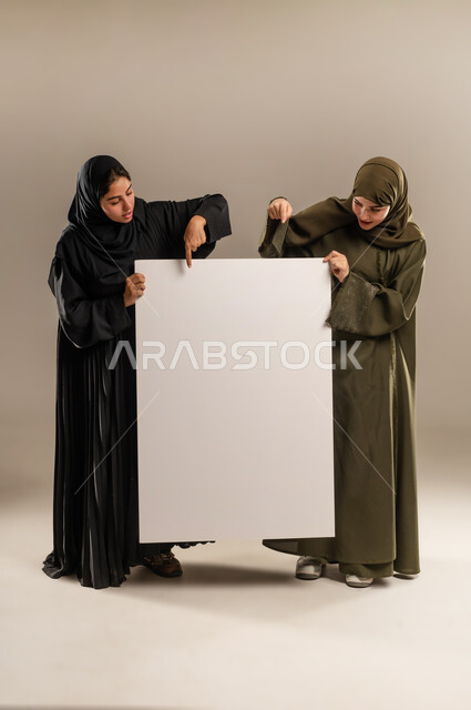Using illustrations and educational tools, blank billboard, pointing the index finger with gestures of pleasure and admiration, advertisements and marketing offers, portrait of two Saudi Arabian Gulf women wearing hijab and abaya holding a large rectangular mockup, full-length body, gray background