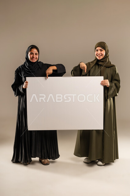 استخدام لوحات توضيحية ووسائل تعليمية، لوح اعلاني فارغ، الإشارة بإصبع السبابة بإيماءات السرور والاعجاب، إعلانات وعروض تسويقية، بورتريه لإمرأتين عربيتين خليجيتين سعوديتين ترتديان الحجاب والعباءة تمسكان موك اب مستطيل كبير، الجسم بالطول الكامل، خلفية رمادية