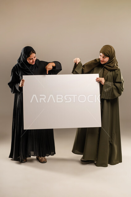 لوح اعلاني فارغ، الإشارة بإصبع السبابة بإيماءات السرور والاعجاب، استخدام لوحات توضيحية ووسائل تعليمية، إعلانات وعروض تسويقية، بورتريه لإمرأتين عربيتين خليجيتين سعوديتين ترتديان الحجاب والعباءة تمسكان موك اب مستطيل كبير، الجسم بالطول الكامل، خلفية رمادية