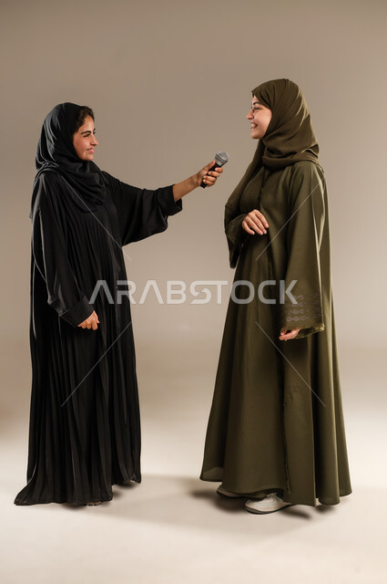 تسجيل المقاطع الصوتية باستخدام الميكرفون، الاستمتاع بالغناء مع الاصدقاء, الدعم الفني للمواهب الغنائية في السعودية, بورتريه لإمرأتين عربيتين خليجيتين سعوديتين ترتديان العباءة والحجاب تقومان بتجربة الغناء المسرحي, صورة الجسم بالكامل, خلفية رمادية