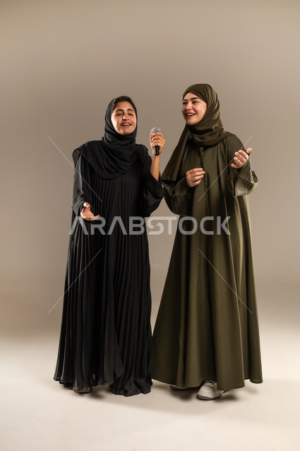 الاستمتاع بالغناء مع الاصدقاء, تسجيل المقاطع الصوتية باستخدام الميكرفون, الدعم الفني للمواهب الغنائية في السعودية, بورتريه لإمرأتين عربيتين خليجيتين سعوديتين ترتديان العباءة والحجاب تقومان بتجربة الغناء المسرحي, صورة الجسم بالكامل, خلفية رمادية