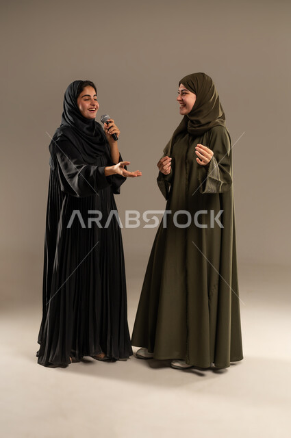 تسجيل المقاطع الصوتية باستخدام الميكرفون، الاستمتاع بالغناء مع الاصدقاء, الدعم الفني للمواهب الغنائية في السعودية, بورتريه لإمرأتين عربيتين خليجيتين سعوديتين ترتديان العباءة والحجاب تقومان بتجربة الغناء المسرحي, صورة الجسم بالكامل, خلفية رمادية