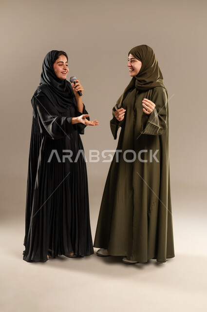 الاستمتاع بالغناء مع الاصدقاء, تسجيل المقاطع الصوتية باستخدام الميكرفون, الدعم الفني للمواهب الغنائية في السعودية, بورتريه لإمرأتين عربيتين خليجيتين سعوديتين ترتديان العباءة والحجاب تقومان بتجربة الغناء المسرحي, صورة الجسم بالكامل, خلفية رمادية