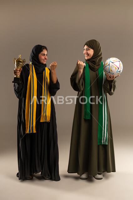 الامساك بالكرة والكأس الذهبي بإيماءات الحماس و الشغف، رفع اليدين بإيماءات الفوز، المباريات والألعاب الرياضية، تعابير الفرح والسرور لانتصار الفريق، بورتريه لإمرأتين عربيتين خليجيتين سعوديتين ترتديان أوشحة تشجيعية لفرق كرة القدم، صورة لكامل الجسم، خلفية رمادية