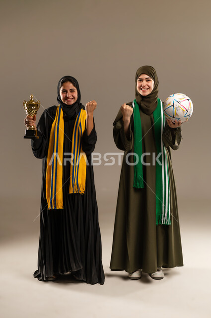 الامساك بالكرة والكأس الذهبي بإيماءات الحماس و الشغف، رفع اليدين بإيماءات الفوز، المباريات والألعاب الرياضية، تعابير الفرح والسرور لانتصار الفريق، بورتريه لإمرأتين عربيتين خليجيتين سعوديتين ترتديان أوشحة تشجيعية لفرق كرة القدم، صورة لكامل الجسم، خلفية رمادية