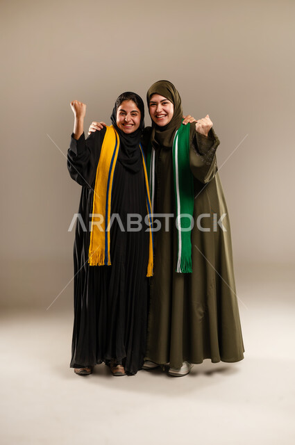 رفع اليدين بإيماءات الفوز، النظر الى الكاميرا بإيماءات الحماس و الشغف، المباريات والألعاب الرياضية، تعابير الفرح  والسرور لانتصار الفريق، بورتريه لإمرأتين عربيتين خليجيتين سعوديتين ترتديان أوشحة تشجيعية لفرق كرة القدم، صورة لكامل الجسم، خلفية رمادية