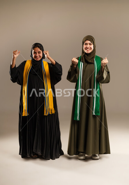 رفع اليدين بإيماءات الفوز، النظر الى الكاميرا بإيماءات الحماس و الشغف، المباريات والألعاب الرياضية، تعابير الفرح  والسرور لانتصار الفريق، بورتريه لإمرأتين عربيتين خليجيتين سعوديتين ترتديان أوشحة تشجيعية لفرق كرة القدم، صورة لكامل الجسم، خلفية رمادية