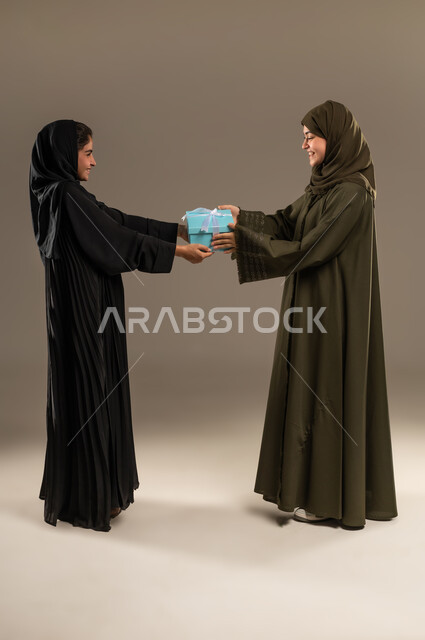 إيماءات الخجل و السرور لتلقي هدية، التعبير عن المحبة والألفة بين الأخوات, تقديم الهدايا والتهنئات، أعياد ومناسبات ومفاجآت سعيدة, بورتريه لإمرأة عربية خليجية سعودية ترتدي العباءة والحجاب تحمل بيدها علبة هدايا تقوم بتقديمها لصديقتها، صورة لكامل الجسم، خلفية رمادية