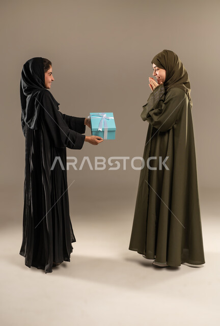 إيماءات الخجل و السرور لتلقي هدية، التعبير عن المحبة والألفة بين الأخوات, تقديم الهدايا والتهنئات، أعياد ومناسبات ومفاجآت سعيدة, بورتريه لإمرأة عربية خليجية سعودية ترتدي العباءة والحجاب تحمل بيدها علبة هدايا تقوم بتقديمها لصديقتها، صورة لكامل الجسم، خلفية رمادية