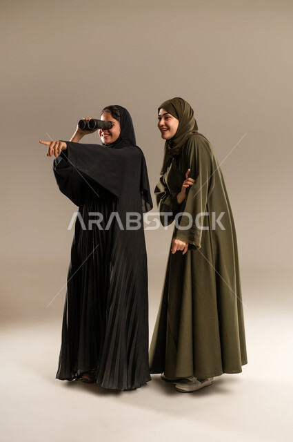 The concept of sharing, exploration and adventure, future ambitions and goals, research using binoculars, portrait of two Saudi Arabian Gulf women sharing a look through binoculars, full-length body image, gray background