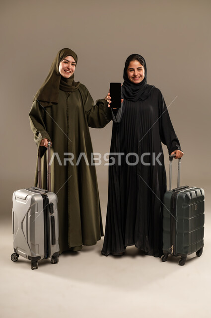 Blank black screen on mobile phone, packing and holding luggage, concept of traveling with friends, portrait of two Saudi Arabian Gulf female friends wearing hijab and abaya getting ready for a fun trip with excited gestures, full body, colorful background