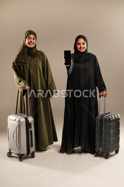 عرض شاشة سوداء فارغة على الهاتف المحمول، تجهيز حقائب الأمتعة والإمساك بها، مفهوم السفر برفقة الأصدقاء، بورتريه لصديقتين عربيتين خليجيتين سعوديتين ترتديان الحجاب والعباءة تقومان بالاستعداد للانطلاق في رحلة ممتعة بإيماءات الحماس، الجسم بالطول الكامل، خلفية ملونة