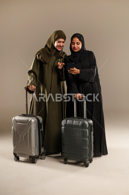 استخدام الهاتف المحمول لمعرفة الوجهه، تجهيز حقائب الأمتعة والإمساك بها، مفهوم السفر برفقة الأصدقاء، بورتريه لصديقتين عربيتين خليجيتين سعوديتين ترتديان الحجاب والعباءة تقومان بالاستعداد للانطلاق في رحلة ممتعة بإيماءات الحماس، الجسم بالطول الكامل، خلفية ملونة
