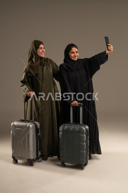 مشاركة الرحلة عبر وسائل التواصل الاجتماعي، تجهيز حقيبة الأمتعة والاستعداد للسفر برفقة الأصدقاء، بورتريه لإمرأتين عربيتين خليجيتين سعوديتين ترتديان العباءة والحجاب تقومان بالتقاط صور السيلفي عبر الهاتف المحمول، الجسم بالطول الكامل، خلفية ملونة