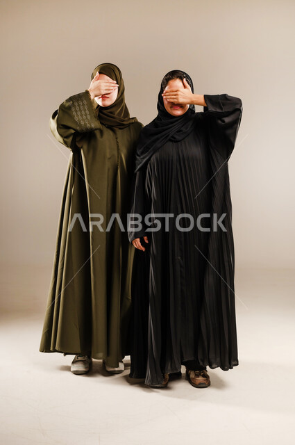 Gestures of shyness and joy, hands over eyes, portrait of two smiling Saudi Arabian Gulf women wearing abaya and hijab closing their eyes, concept of body language and gestures, full body image, gray background