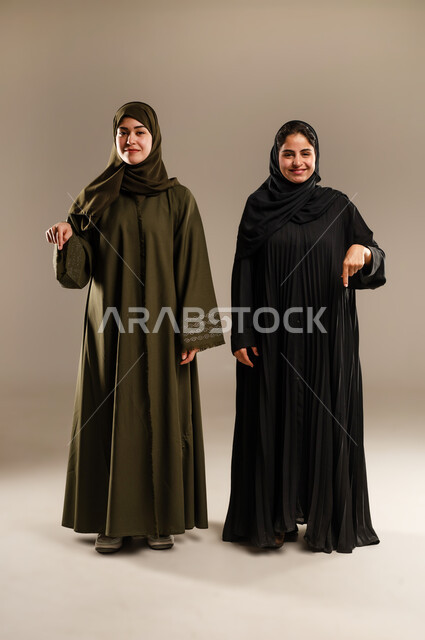 حركات تدل على تحديد المكان، الإشارة إلى شيء ما بأصبع السبابة، مفهوم لغة الجسد والحركات والإشارات، بورتريه لإمرأتين عربيتين سعوديتين خليجيتين ترتديان الحجاب والعباءة تشيران الى الاسفل، صورة الجسم بالطول الكامل، خلفية رمادية