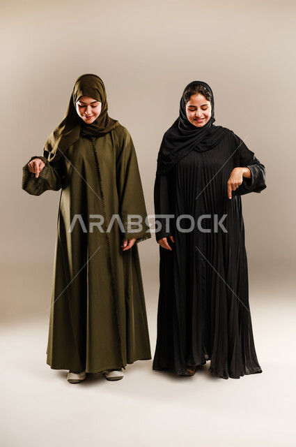 حركات تدل على تحديد المكان، الإشارة إلى شيء ما بأصبع السبابة، مفهوم لغة الجسد والحركات والإشارات، بورتريه لإمرأتين عربيتين سعوديتين خليجيتين ترتديان الحجاب والعباءة تشيران الى الاسفل، صورة الجسم بالطول الكامل، خلفية رمادية