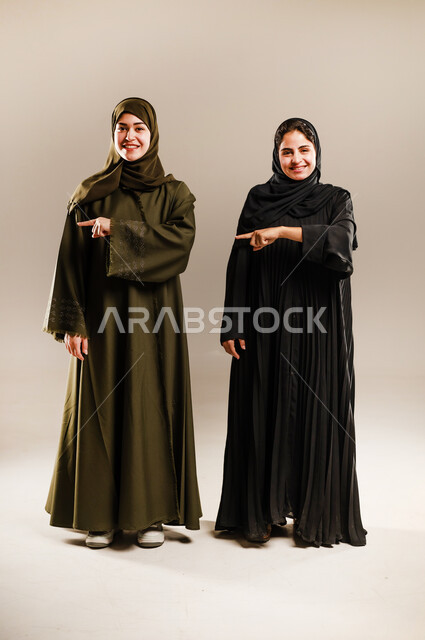 الإشارة إلى شيء ما بأصبع السبابة، حركات تدل على تحديد المكان، مفهوم لغة الجسد والحركات والإشارات، بورتريه لإمرأتين عربيتين سعوديتين خليجيتين ترتديان الحجاب والعباءة تشيران الى الجانب، صورة الجسم بالطول الكامل، خلفية رمادية
