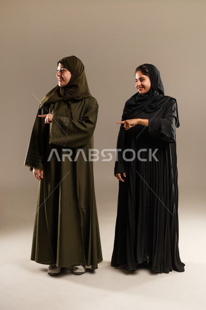 الإشارة إلى شيء ما بأصبع السبابة، حركات تدل على تحديد المكان، مفهوم لغة الجسد والحركات والإشارات، بورتريه لإمرأتين عربيتين سعوديتين خليجيتين ترتديان الحجاب والعباءة تشيران الى الجانب، صورة الجسم بالطول الكامل، خلفية رمادية