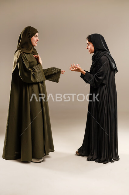 مفهوم النقاش الحاد والمواجهة بين الأخوات، بورتريه لإمرأتين عربيتين خليجيتين سعوديتين ترتديان الحجاب والعباءة تقفان مقابل بعضهما وتتجادلان بإيماءات الغضب وإلقاء اللوم، تعابير التعصب والتوتر الانفعالي، الجسم بالطول الكامل، خلفية رمادية