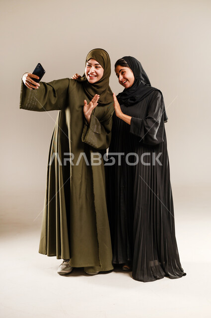 استخدام الأجهزة التقنية المتطورة، توثيق اللحظات السعيدة، بورتريه لشابة عربية خليجية سعودية محجبة ترتدي العباءة والحجاب تقوم بالتقاط صور السيلفي التذكارية مع صديقتها بإيماءات السعادة والسرور، صورة الجسم بالكامل، خلفية رمادية