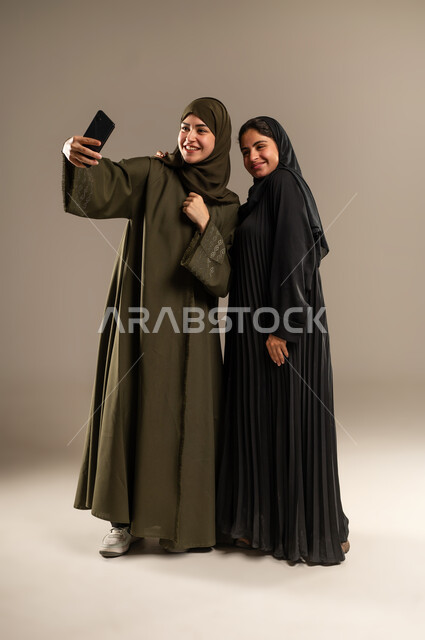Using advanced technical devices, documenting happy moments, a portrait of a veiled Saudi Arabian Gulf young woman wearing an abaya and hijab, taking commemorative selfies with her friend, with gestures of happiness and pleasure, a full body image, a gray background