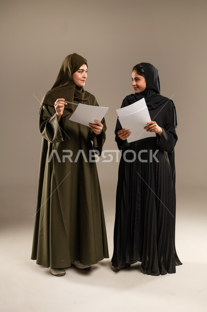 مناقشة الأعمال مع زملاء العمل، المشاركة والتعاون في إنجاز العمل، ايماءات الفرح والسعادة, بورتريه لإمرأتين عربيتين سعوديتين خليجيتين ترتديان الحجاب والعباءة تمسكان مجموعة من الاوراق والوثائق، صورة الجسم بالطول الكامل، خلفية رمادية