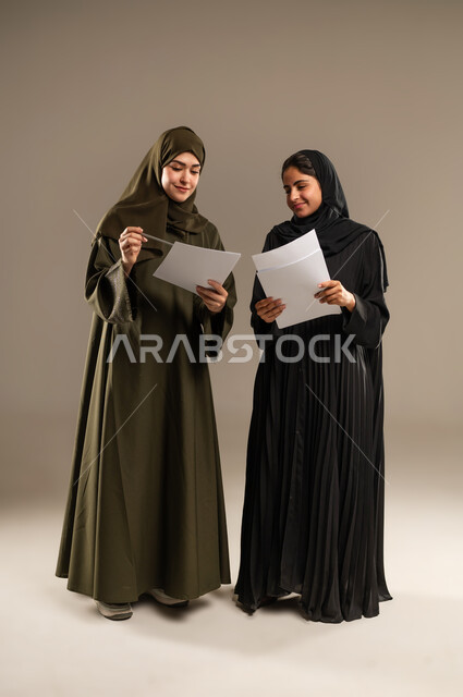 Discussing work with colleagues, sharing and collaborating in completing work, gestures of joy and happiness, portrait of two Saudi Arabian Gulf women wearing hijab and abaya holding a group of papers and documents, full body image, gray background