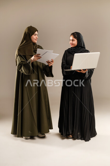الانتباه للملاحظات وتطبيقها، تبادل الأفكار والخبرات لتطوير استراتيجيات العمل، بورتريه لإمرأتين عربيتين سعوديتين خليجيتين ترتديان الحجاب والعباءة تقومان بتطوير الخطط وحل المشكلات لتحقيق أهداف الشركة باستخدام الحاسب المحمول، الجسم بالطول الكامل، خلفيه رمادية