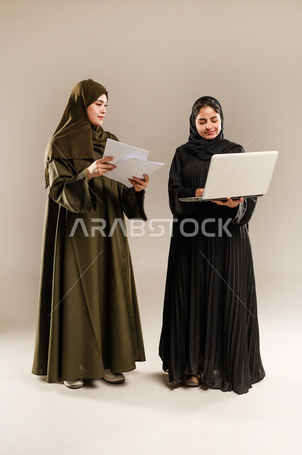 Paying attention to feedback and implementing it, exchanging ideas and experiences to develop business strategies, portrait of two Saudi Arabian Gulf women wearing hijab and abaya developing plans and solving problems to achieve company goals using laptop, full body, gray background