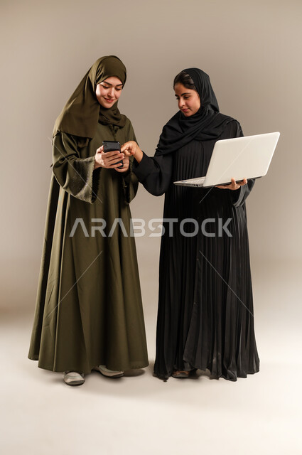 تبادل الأفكار والخبرات لتطوير استراتيجيات العمل، بورتريه لإمرأتين عربيتين سعوديتين خليجيتين ترتديان الحجاب والعباءة تقومان بتطوير الخطط وحل المشكلات لتحقيق أهداف الشركة باستخدام الهاتف المحول والحاسب، الانتباه للملاحظات وتطبيقها، الجسم بالطول الكامل، خلفيه رمادية