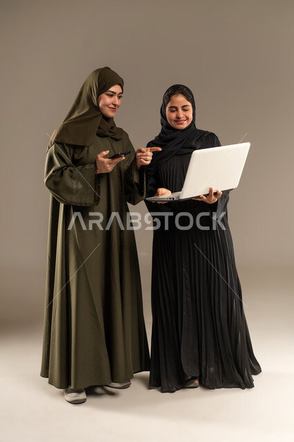 الانتباه للملاحظات وتطبيقها، تبادل الأفكار والخبرات لتطوير استراتيجيات العمل، بورتريه لإمرأتين عربيتين سعوديتين خليجيتين ترتديان الحجاب والعباءة تقومان بتطوير الخطط وحل المشكلات لتحقيق أهداف الشركة باستخدام الهاتف المحول والحاسب، الجسم بالطول الكامل، خلفيه رمادية