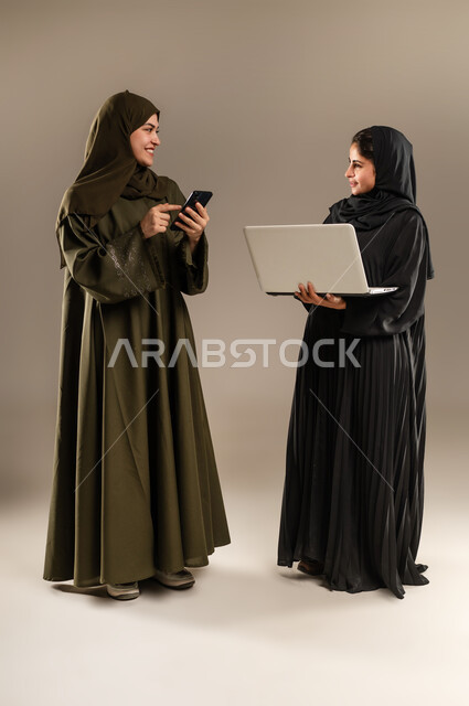 Paying attention to feedback and implementing it, exchanging ideas and experiences to develop business strategies, portrait of two Saudi Arabian Gulf women wearing hijab and abaya developing plans and solving problems to achieve company goals using mobile phone and computer, full body, gray background