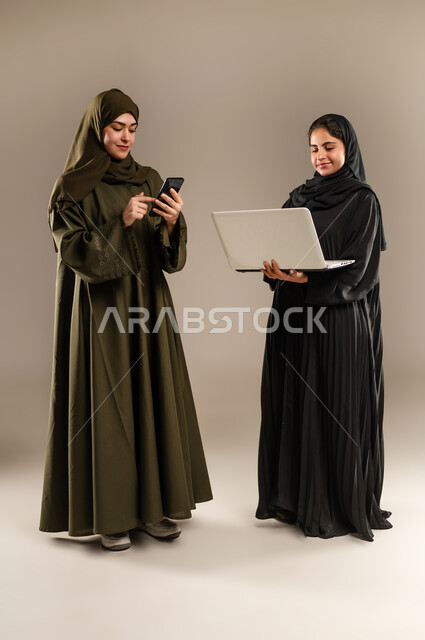 Paying attention to feedback and implementing it, exchanging ideas and experiences to develop business strategies, portrait of two Saudi Arabian Gulf women wearing hijab and abaya developing plans and solving problems to achieve company goals using mobile phone and computer, full body, gray background