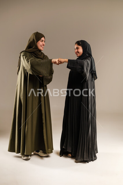 الحفاظ على المسافات بين الاشخاص، اتباع احترازات السلامة، بورتريه لإمرأتين عربيتين خليجيتين سعودتين محجبتين تقومان بالتصافح بقبضة اليد، الالتزام بإجراءات الوقاية من فايروس كورونا، صورة لكامل الجسم، خلفية رمادية