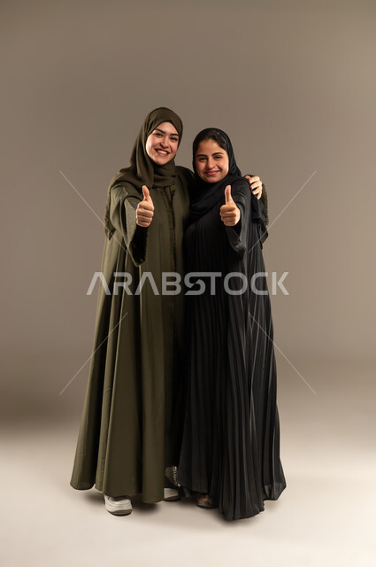 الاتفاق على أمر معين، شابة تعانق اختها، القبول والرضا بالنتيجة، ايماءات الاعجاب والموافقة،  بورتريه لإمرأتين عربيتين خليجيتين سعوديتين ترتديان الحجاب والعباءة ترفعان اصبع الابهام للأعلى بعلامة الجودة، صورة لكامل الجسم، خلفية رمادية