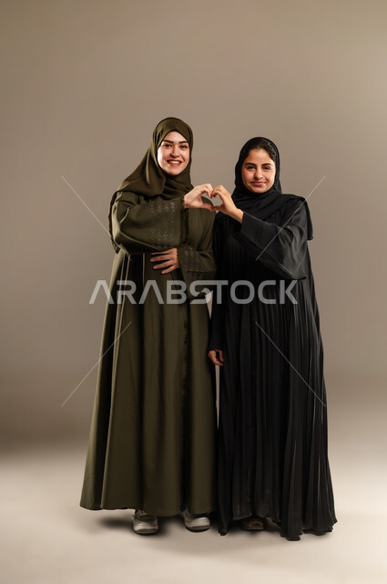 إشارة يدين على شكل قلب حب بايماءات وجه ويدين تدل على الشعور بالحب، بورتريه لإمرأتين عربيتين خليجيتين سعوديتين ترتديان العباءة والحجاب تقفان بتعابير السرور، العلاقة الايجابية الاخوية والحب والمودة، صورة الجسم بالطول الكامل، خلفية رمادية