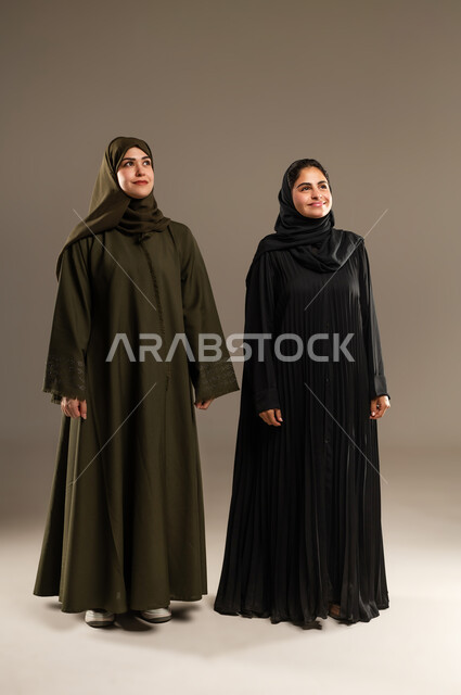 Standing straight and looking at something with gestures of joy, portrait of two smiling Saudi Arabian Gulf women wearing abaya and hijab with expressions of happiness and pleasure, full body portrait, gray background
