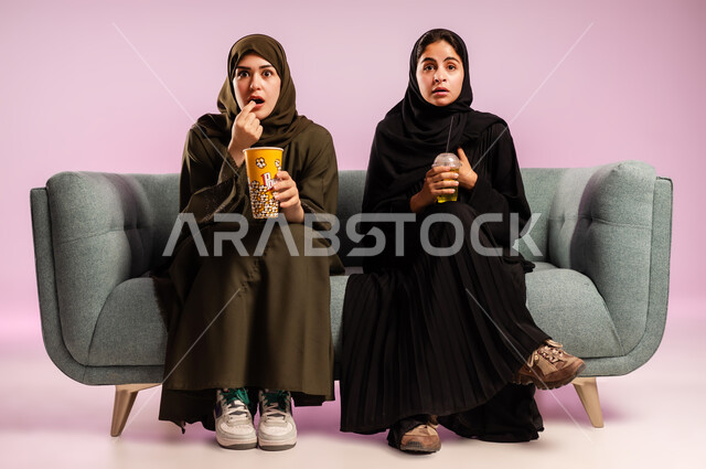قضاء وقت ممتع في السينما، تناول الفوشار والعصير اثناء متابعة الأفلام المسلية، بورتريه لإمرأتين عربيتين خليجيتين سعوديتين ترتديان الحجاب والعباءة تجلسان على كنبة تقومان بأنشطة ترفيهيه في عطلة نهاية الإسبوع، خلفية ملونة