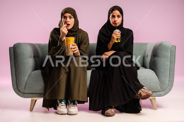 تناول الفوشار والعصير اثناء متابعة الأفلام المسلية، بورتريه لإمرأتين عربيتين خليجيتين سعوديتين ترتديان الحجاب والعباءة تجلسان على كنبة تقومان بأنشطة ترفيهيه في عطلة نهاية الإسبوع، قضاء وقت ممتع في السينما، خلفية ملونة