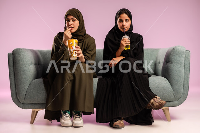 قضاء وقت ممتع في السينما، تناول الفوشار والعصير اثناء متابعة الأفلام المسلية، بورتريه لإمرأتين عربيتين خليجيتين سعوديتين ترتديان الحجاب والعباءة تجلسان على كنبة تقومان بأنشطة ترفيهيه في عطلة نهاية الإسبوع، خلفية ملونة