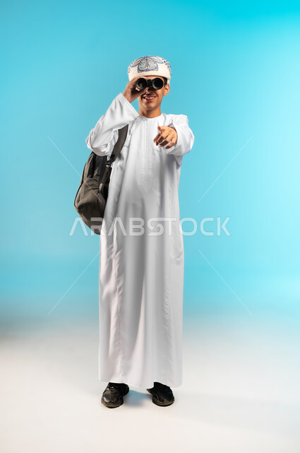 Using binoculars to detect and bring distant objects closer, love of adventure and exploration, future planning for goals and ambitions, portrait of an Arab Gulf Omani boy wearing a dishdasha and a sleeve looking through a binocular view (telescope) with gestures of joy and happiness, full-length body image, blue background.