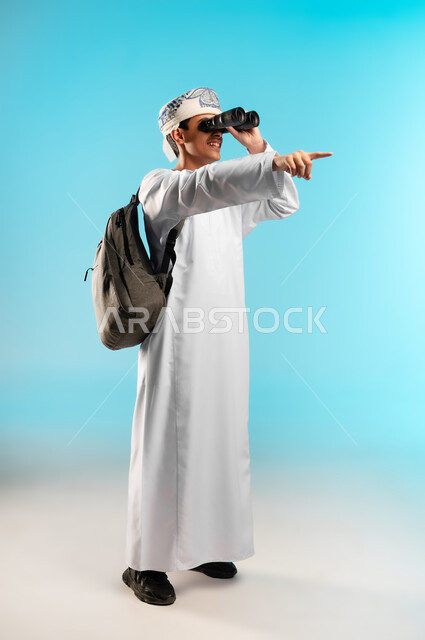 التخطيط المستقبلي للأهداف والطموحات،  حب المغامرة والاستكشاف، بورتريه لولد عربي خليجي عماني يرتدي الدشداشة والكمه ينظر من خلال المنظور ثنائي العينين (التلسكوب) بإيماءات الفرح والسرور، رؤية الاشياء البعيدة عبر المنظار، صورة الجسم بالطول الكامل، خلفية زرقاء .
