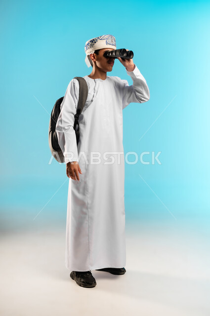 رؤية الاشياء البعيدة عبر المنظار، حب المغامرة والاستكشاف، بورتريه لولد عربي خليجي عماني يرتدي الدشداشة والكمه ينظر من خلال المنظور ثنائي العينين (التلسكوب) بإيماءات الفرح والسرور، التخطيط المستقبلي للأهداف والطموحات، صورة الجسم بالطول الكامل، خلفية زرقاء .