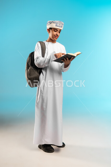 المناهج العلمية التعليمية، الاجتهاد والتفوق ومتابعة الدراسة ، مفهوم مراجعة الدروس وحل الواجبات، الاستعداد للذهاب إلى المدرسة، بورتريه لولد عربي خليجي عماني يرتدي الدشداشة والكمه يحمل على ظهرة حقيبة مدرسية وبيده كتاب ، النظر الى الاعلى بتعابير السعادة ، صورة الجسم بالطول الكامل, خلفية زرقاء .